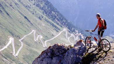 Un ciclista di montagna si trova in cima a una vetta, davanti a lui una vista mozzafiato sui passi serpeggianti di Vinschgau in Alto Adige.