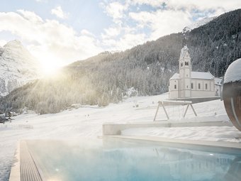 Heated outdoor pool of Hotel Bella Vista at sunset with sunbeams shining through the trees, illuminating the steam above the water in the snow-covered South Tyrolean landscape.