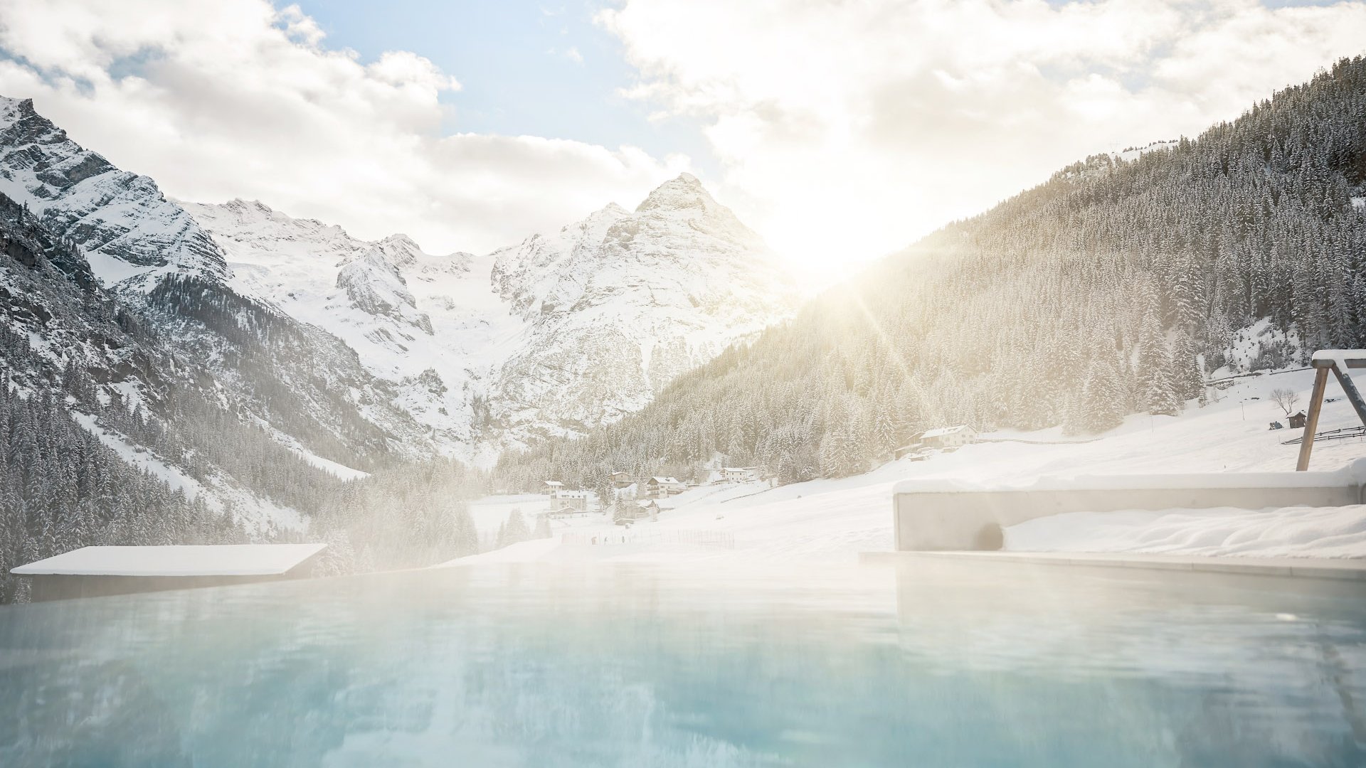 Wintry swimming pleasure in the heated outdoor pool of Hotel Bella Vista with a view of snow-covered trees and mountain scenery at sunset.