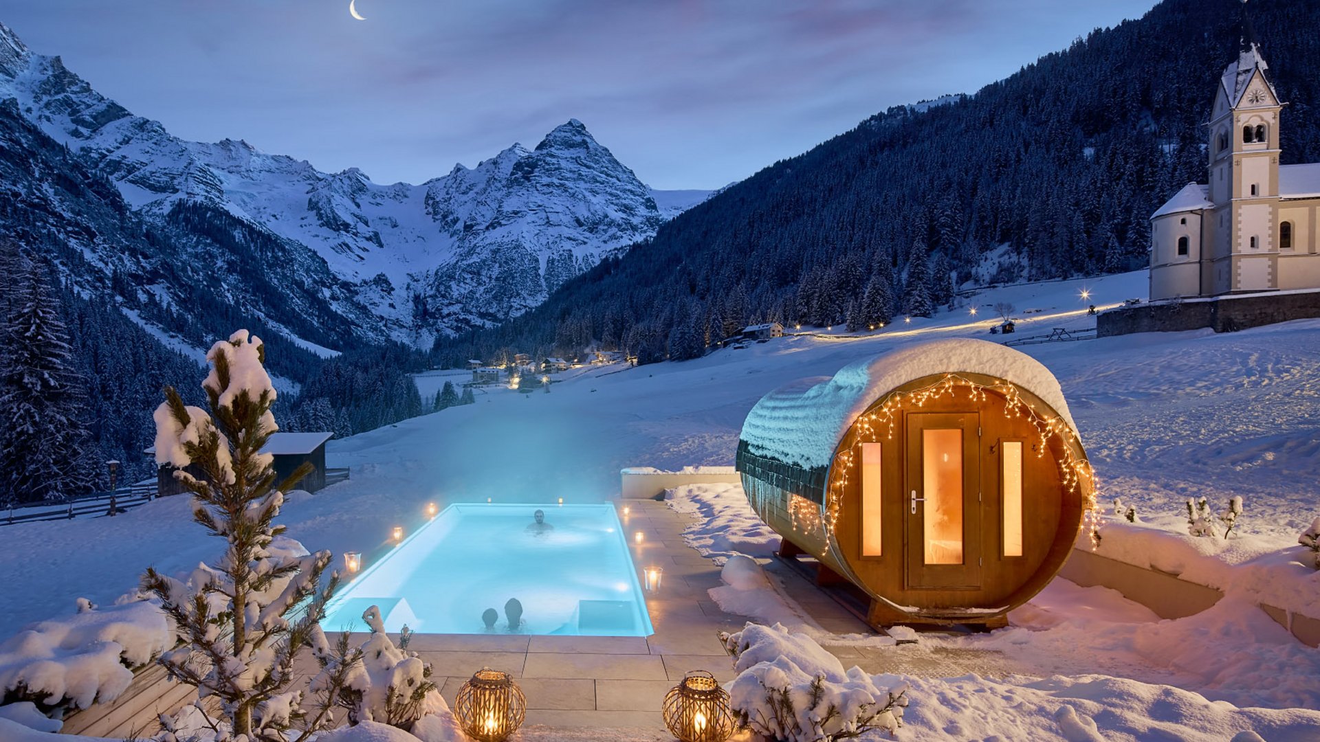 Night view of the heated outdoor pool and adjacent sauna at Hotel Bella Vista, with a chapel in the background, nestled in South Tyrol's snow-covered mountain landscape.