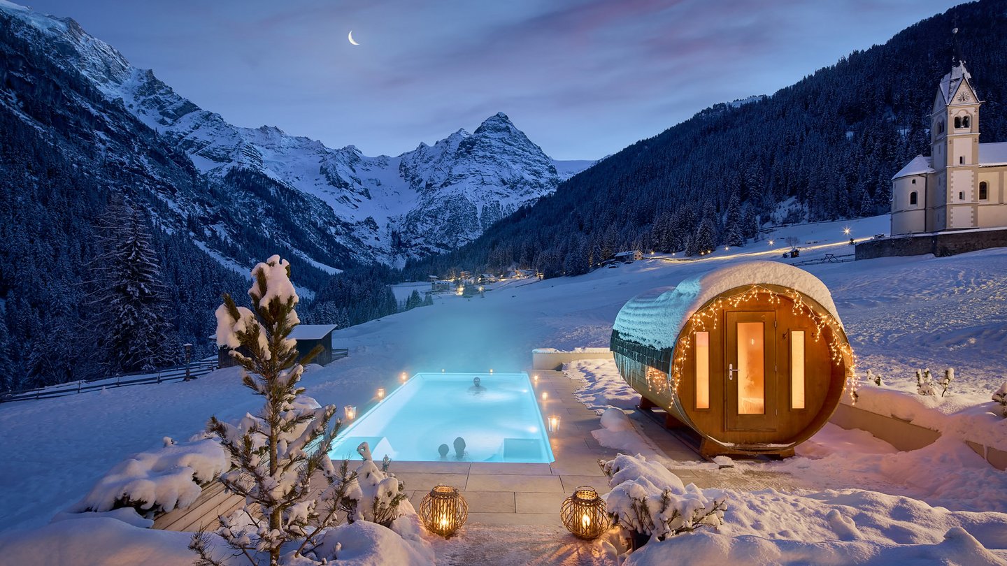 Nachtansicht des beheizten Außenpools und der angrenzenden Sauna am Hotel Bella Vista mit einer Kapelle im Hintergrund, eingebettet in die schneebedeckte Berglandschaft Südtirols.