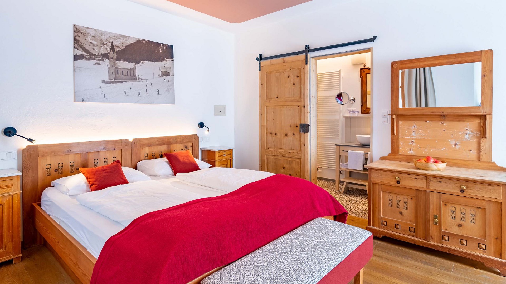 A cozy room at Hotel Bella Vista featuring traditional South Tyrolean wooden furniture, red accent bedding, and an inviting atmosphere for a restful night.