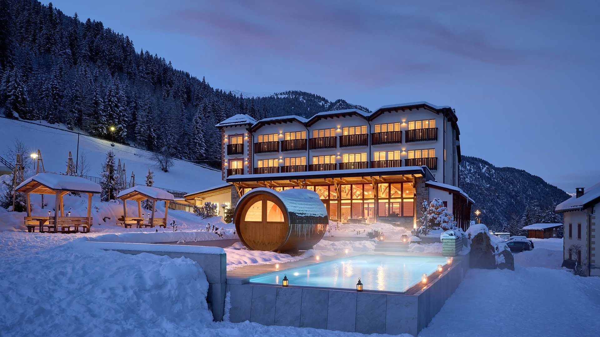 Evening view of Hotel Bella Vista in South Tyrol with lit windows and a steaming outdoor pool, surrounded by a snowy mountain landscape.